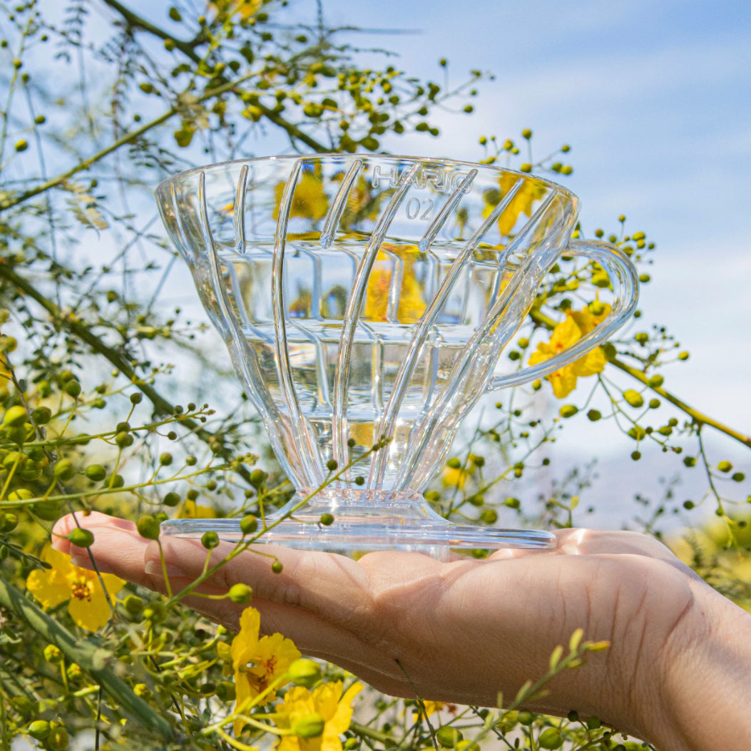 Clear, plastic coffee pour over being held in a hand with yellow flowers and sky in the background. Available through the Awesome Coffee Club.