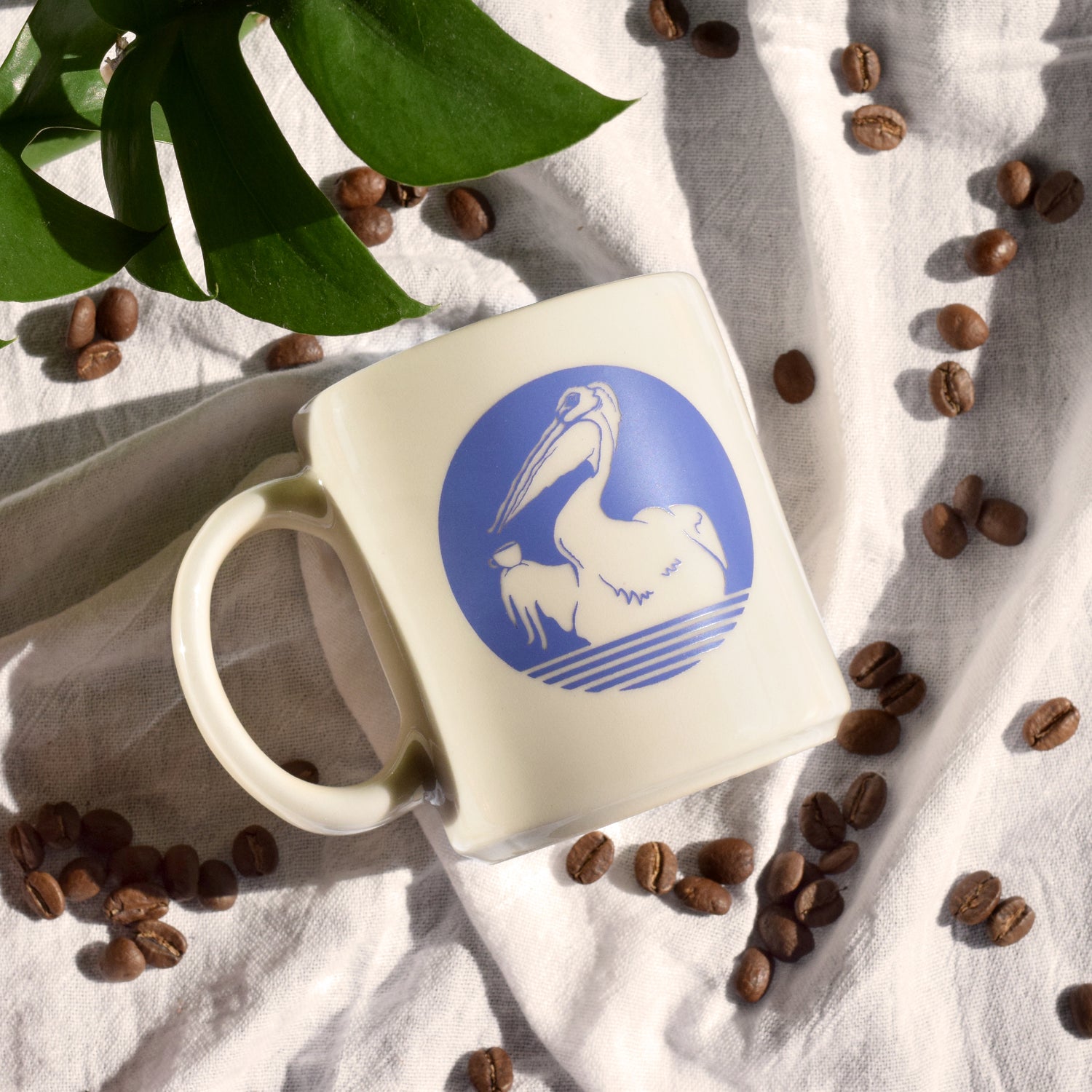 White cafe mug with the outline of a pelican holding a cup of coffee from the Awesome Coffee Club logo. The mug is laid on a sheet with a plant in the corner and coffee beans surrounding it. 