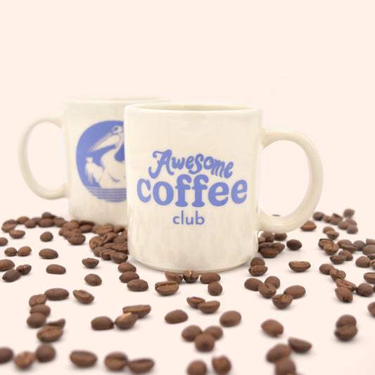 White cafe mug with the Awesome Coffee Club logo in purple written on the front. Behind the mug is a second mug with the outline of a pelican holding a cup, showing the back of the mug. Coffee beans surround both mugs. 